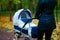 Young caucasian woman is pushing white baby carriage while she walking at autumn park with beautiful yellow leaves