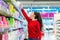 A young Caucasian woman reaches to the top shelf for a pack of sanitary pads. Side view. Corridor of shop on the