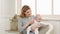 A young caucasian woman sits in a chair by the window and holds a newborn baby girl. Home clothes.