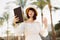 Young caucasian woman sitting on the background of palm trees, taking photo by tablet