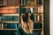 Young caucasian woman standing near bookshelf in library and choosing book for reading