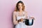 Young caucasian woman wearing apron holding cooking pot sticking tongue out happy with funny expression