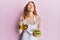 Young caucasian woman wearing apron holding olive oil can and salad looking at the camera blowing a kiss being lovely and sexy