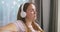 A young Caucasian woman wearing wireless headphones sits on a couch at home