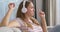 A young Caucasian woman wearing wireless headphones sits on a couch at home