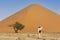 Young caucasic traveler standing near orange sand dune with her hand on the hat. Travel desert concept