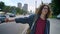 Young charming student in glasses with long wavy hair waiting for taxi and standing near road with cars, urban modern