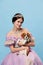 Young charming woman in lilac color vintage dress as royal person, princess holding small King Charles spaniel dog on