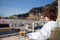 Young charming woman wearing in white terry robe is drinking coffee and relaxing in deck chair on balcony of hotel in Monaco.