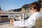 Young charming woman wearing in white terry robe is drinking coffee and relaxing in deck chair on balcony of hotel in Monaco.
