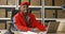 Young cheerful African American postman in red iniform and cap sitting at desk in postal office store and working at