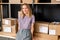 Young cheerful female worker of online shop office standing against large shelf