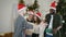 Young cheerful friends in Santa hats celebrating Christmas are standing before christmas tree, holding present boxes and
