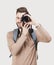 Young cheerful man photographer takes images with digital camera. Isolated on gray background. Studio shot.