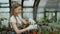 Young cheerful woman in apron and gloves gardening plants and loosen ground in flower in greenhouse