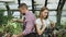 Young cheerful woman in apron and gloves loosen ground in flower and chatting his boyfriend in greenhouse