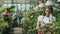 Young cheerful woman in apron and gloves talking phone while gardening plants and loosen ground in flower in greenhouse