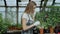 Young cheerful woman in apron and gloves talking phone while gardening plants and loosen ground in flower in greenhouse