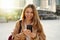 Young cheerful woman using cell phone and texting message on city street. Happy girl holding smart phone in hands and smiling.