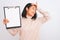 Young chinese inspector woman holding clipboard standing over isolated white background stressed with hand on head, shocked with