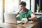 Young cold sick businessman in green t-shirt sitting at work, sneezing and cleaning his nose with tissue napkin