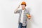 Young concerned man in protective orange hardhat holding toy saw and beating himself on head with toy hammer isolated on