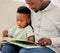 Young content African american father reading a book with his son sitting on the couch at home. Little boy enjoying a