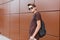 Young cool hipster man in dark stylish sunglasses in trendy summer clothes with a black backpack poses near the brown wall