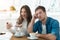 Young couple beautiful woman communicates via smartphone and handsome man looks annoyed while drinking coffee in cafe ,