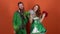 Young couple in costumes celebrating saint patrick`s day isolated on orange wall shaking pom poms