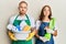 Young couple of girlfriend and boyfriend wearing apron holding products and cleaning spray puffing cheeks with funny face