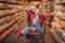 Young couple in grocery store. Woman sit in trolley and have fun. Man stand behind and point on shelf with products