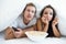 Young couple handsome man and beautiful woman watching TV in bed eating pop corn looking fascinated