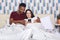 Young couple lying in bed, female iholding mobile phone, husband hugs his wife, watching film online, having rest together,