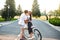 Young couple of romantic people on road on sunset. Pretty girl with long curly hair in hat and long skirt holds a bike