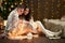 Young couple sit on floor in dark wooden interior with lights. Romantic evening and love concept. New year holiday. Christmas ligh