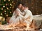 Young couple sit on floor in dark wooden interior with lights. Romantic evening and love concept. New year holiday. Christmas ligh