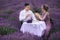 Young couple are sitting on chairs at table and drink coffee or tea in lavender field. woman in purple dress and flower`s wreath