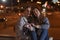 Young couple is sitting on the pavement on evening city background with garland in their hands. Night city lights