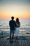 Young couple watching the sunrise at the beach, back view, seashore summer beach at yellow blue morning horizon sea