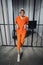 The young criminal is leaning against the bars of the cell in an orange robe. Women`s maximum security prison