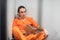 Young criminal in a solitary cell reads a book sitting on a bunk in an orange prison uniform. Photo through the bars.