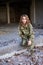 Young curly blond military woman, wearing ukrainian military uniform, sitting on pavement edge, smiling. Full-length portrait of
