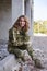 Young curly blond military woman, wearing ukrainian military uniform, sitting on pavement edge, smiling. Full-length portrait of