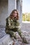 Young curly blond military woman, wearing ukrainian military uniform, sitting on pavement edge, smiling. Full-length portrait of