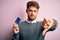 Young customer man with beard holding credit card for payment over pink background with angry face, negative sign showing dislike