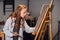 A young cute artist girl is learning to draw in an art workshop. Draws on an easel with oil paints and a brush.