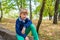 A young and cute boy in green jeans sits on a curb in a park with his hand on his knee