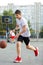 A young cute boy plays basketball at the outdoor streetball court on a sunny summer day. Teenager player in action dribbling