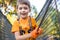 Young cute child boy in summer clothing, safety harness and helmet attached with carbine to cable moves slowly along rope way on